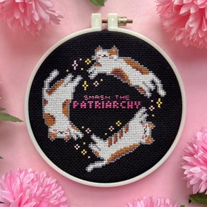 Può includere: Un telaio per punto croce con tre gatti su sfondo nero. I gatti sono bianchi con macchie marroni. Il testo "SMASH THE PATRIARCHY" è ricamato in rosa. Il telaio è circondato da fiori rosa.