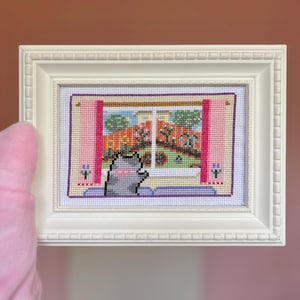 May include: A white-framed cross-stitch picture showing a gray cat gazing out of a window. The window reveals a colourful garden scene with flowers and buildings. The frame has a decorative border, and the artwork is set against a pink background.