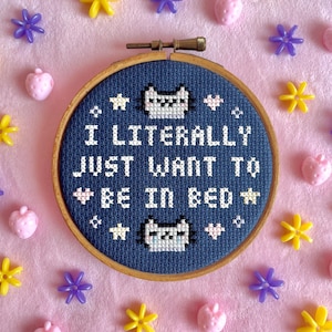 May include: A cross-stitch hoop with the text "I LITERALLY JUST WANT TO BE IN BED" in white thread on a navy blue background. The design includes cat faces and stars. The hoop is surrounded by pink, yellow, and purple decorative elements.