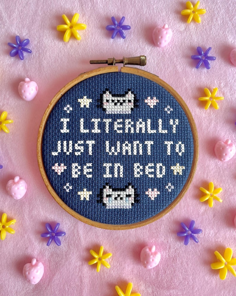 May include: A cross-stitch hoop with the text "I LITERALLY JUST WANT TO BE IN BED" in white thread on a navy blue background. The design includes cat faces and stars. The hoop is surrounded by pink, yellow, and purple decorative elements.