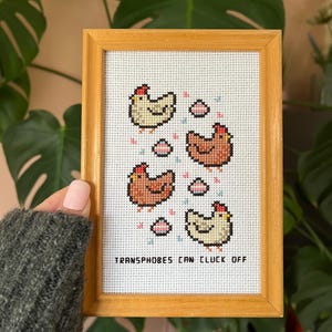 May include: A framed cross-stitch featuring four chickens in shades of brown and cream, with small pink and blue accents. The text "TRANSPHOBES CAN CLUCK OFF" is stitched below the chickens. The frame is made of light-colored wood.