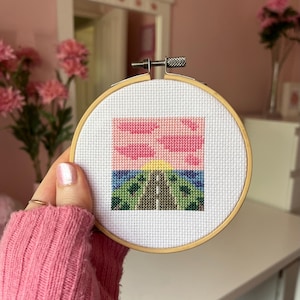Puede incluir: Un paisaje de punto de cruz en un bastidor de madera. La escena representa un camino que conduce a una puesta de sol con tonos rosas y amarillos, y árboles verdes. El bastidor lo sostiene una persona que lleva un jersey rosa.