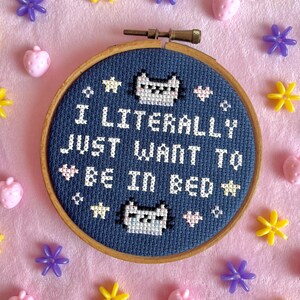 May include: A cross-stitch embroidery hoop with the text "I LITERALLY JUST WANT TO BE IN BED" in white on a navy blue background. The design includes pixelated cat faces and stars. The hoop is surrounded by pink, yellow, and purple decorative elements.