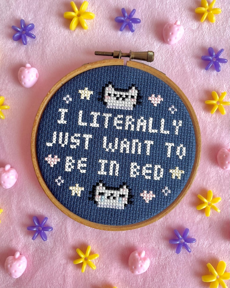 May include: A cross-stitch embroidery hoop with the text "I LITERALLY JUST WANT TO BE IN BED" in white on a navy blue background. The design includes pixelated cat faces and stars. The hoop is surrounded by pink, yellow, and purple decorative elements.