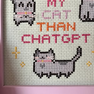 Pode incluir: Arte em ponto cruz em uma moldura rosa. A obra apresenta gatos cinzentos pixelados com coleiras rosa e o texto "I love my cat than ChatGPT" em rosa e laranja. Detalhes de estrelas douradas tamb&eacute;m est&atilde;o inclu&iacute;dos.