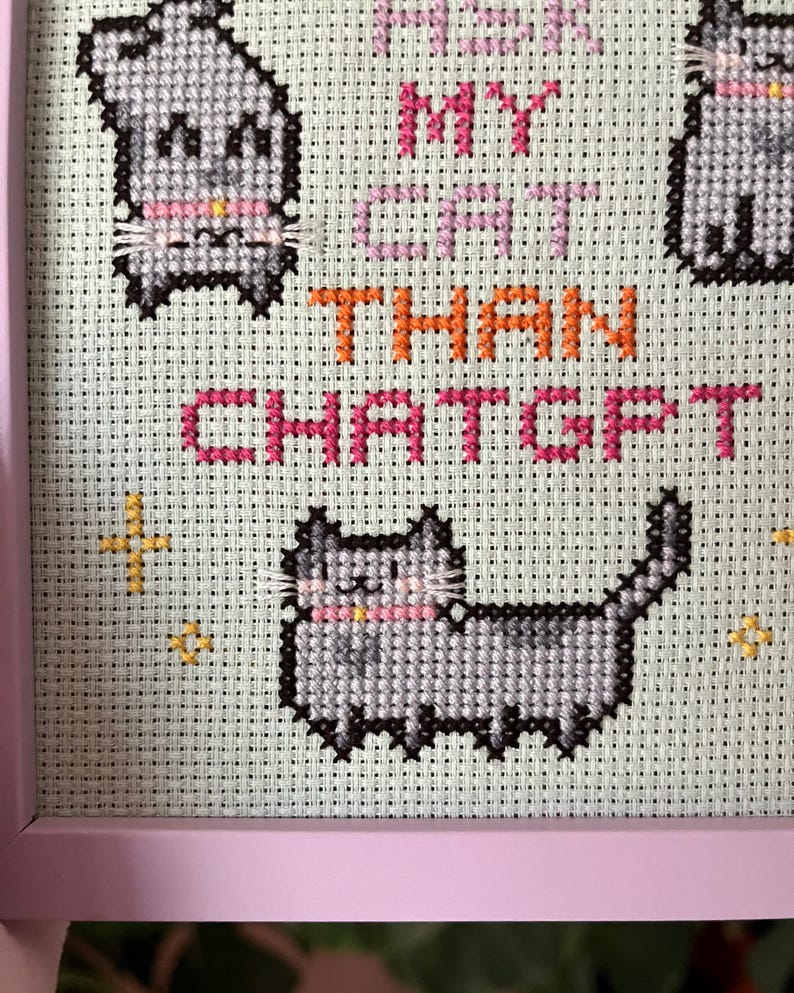 Pode incluir: Arte em ponto cruz em uma moldura rosa. A obra apresenta gatos cinzentos pixelados com coleiras rosa e o texto "I love my cat than ChatGPT" em rosa e laranja. Detalhes de estrelas douradas tamb&eacute;m est&atilde;o inclu&iacute;dos.