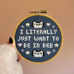 May include: A cross-stitch embroidery hoop with the text "I LITERALLY JUST WANT TO BE IN BED" in white. The design includes cat faces, stars, and hearts on a navy blue background, set in a wooden hoop.