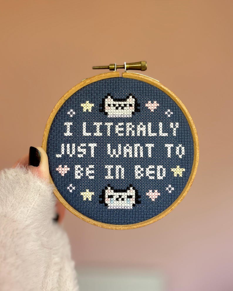 May include: A cross-stitch embroidery hoop with the text "I LITERALLY JUST WANT TO BE IN BED" in white. The design includes cat faces, stars, and hearts on a navy blue background, set in a wooden hoop.