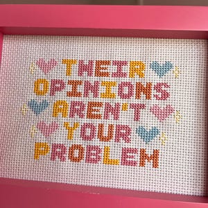 Puede incluir: Un bordado en punto de cruz enmarcado en rosa con el texto "THEIR OPINIONS AREN'T YOUR PROBLEM". Las palabras est&aacute;n bordadas en varios colores sobre tela blanca. Tambi&eacute;n hay corazones y estrellas decorativas bordadas.