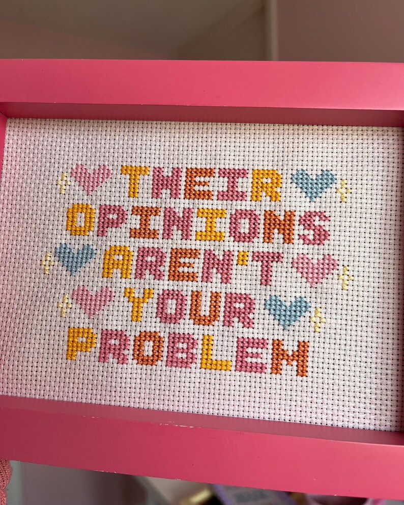 Puede incluir: Un bordado en punto de cruz enmarcado en rosa con el texto "THEIR OPINIONS AREN'T YOUR PROBLEM". Las palabras est&aacute;n bordadas en varios colores sobre tela blanca. Tambi&eacute;n hay corazones y estrellas decorativas bordadas.