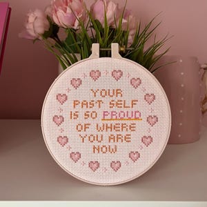 May include: A round, cross-stitched embroidery hoop with the text "YOUR PAST SELF IS SO PROUD OF WHERE YOU ARE NOW" in brown and pink thread. The design includes pink hearts around the edge. The hoop is displayed in front of pink flowers.