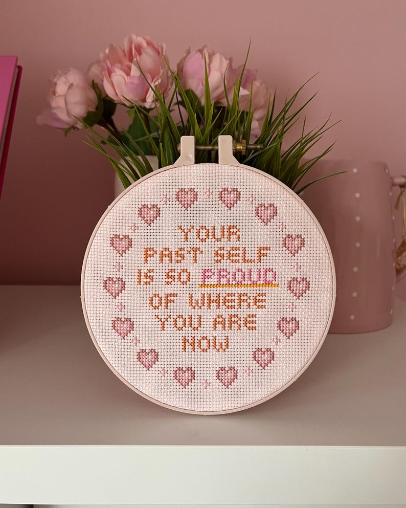 K&ouml;nnte beinhalten: Ein runder Kreuzstich-Stickrahmen mit dem Text "YOUR PAST SELF IS SO PROUD OF WHERE YOU ARE NOW" in braunem und rosa Garn. Das Design umfasst rosa Herzen am Rand. Der Rahmen wird vor rosa Blumen pr&auml;sentiert.