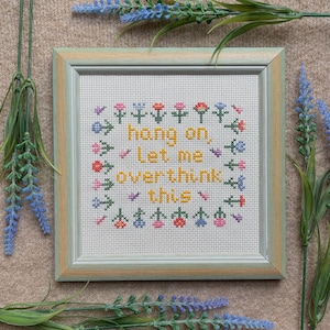 May include: A framed cross-stitch with the words "hang on, let me overthink this" in yellow. The text is surrounded by a floral wreath in various colours. The frame is light green and the background is a neutral colour.
