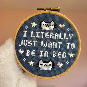 May include: A cross-stitch embroidery hoop with the text "I LITERALLY JUST WANT TO BE IN BED" in white. The design includes cat faces, stars, and hearts on a navy blue background, set in a wooden hoop.