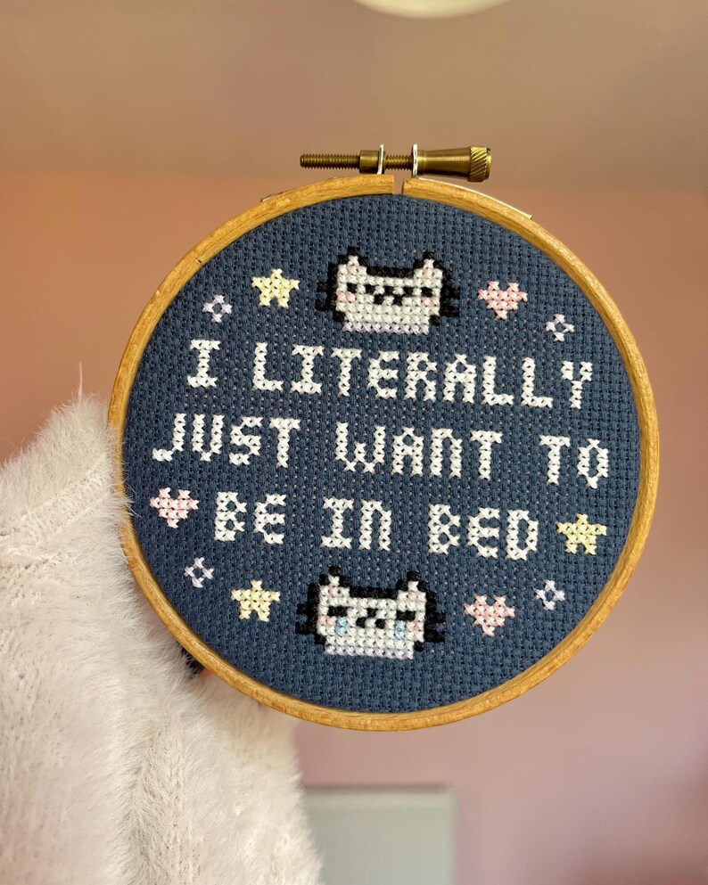 May include: A cross-stitch embroidery hoop with the text "I LITERALLY JUST WANT TO BE IN BED" in white. The design includes cat faces, stars, and hearts on a navy blue background, set in a wooden hoop.