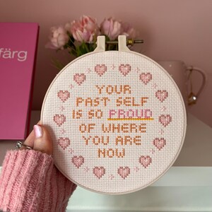 K&ouml;nnte beinhalten: Ein Kreuzstich-Stickrahmen mit dem Text "YOUR PAST SELF IS SO PROUD OF WHERE YOU ARE NOW" in orangefarbenem und wei&szlig;em Garn. Das Design umfasst rosa Herzakzente um den Rand des Reifens.