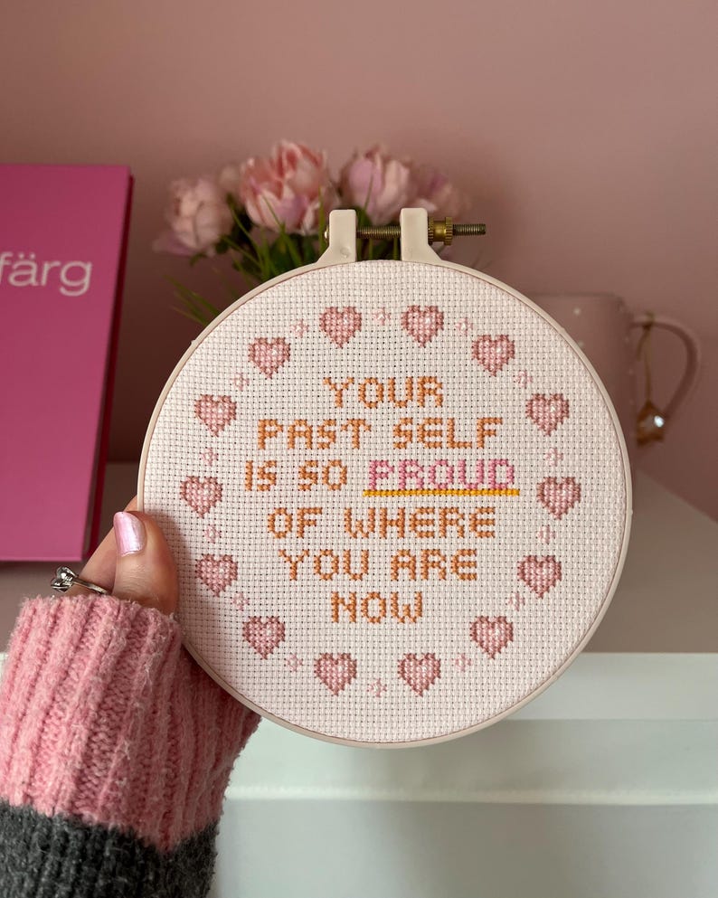 K&ouml;nnte beinhalten: Ein Kreuzstich-Stickrahmen mit dem Text "YOUR PAST SELF IS SO PROUD OF WHERE YOU ARE NOW" in orangefarbenem und wei&szlig;em Garn. Das Design umfasst rosa Herzakzente um den Rand des Reifens.