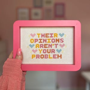 Puede incluir: Un punto de cruz enmarcado en rosa con el texto "THEIR OPINIONS AREN'T YOUR PROBLEM" en letras coloridas y acentos de coraz&oacute;n. El punto de cruz est&aacute; sostenido por una persona que lleva un su&eacute;ter rosa.
