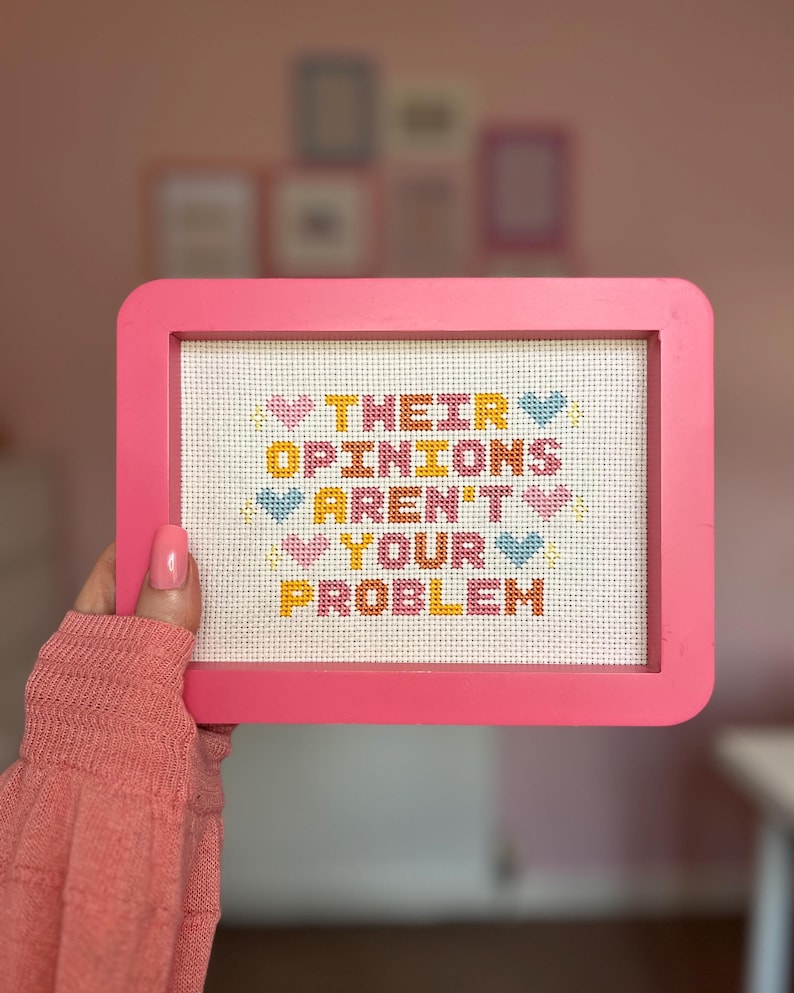 Puede incluir: Un punto de cruz enmarcado en rosa con el texto "THEIR OPINIONS AREN'T YOUR PROBLEM" en letras coloridas y acentos de coraz&oacute;n. El punto de cruz est&aacute; sostenido por una persona que lleva un su&eacute;ter rosa.