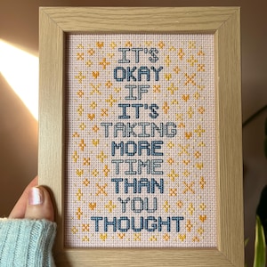 Può includere: Un ricamo a punto croce incorniciato con uno sfondo rosa chiaro e ricami gialli e blu. Il testo dice "It's okay if it's taking more time than you thought."