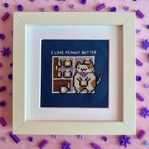 May include: A square white frame containing a cross-stitch embroidery on dark blue fabric. The design features a calico cat holding a jar, standing next to a brown cupboard with shelves. The text "I LOVE PEANUT BUTTER" is stitched in white.