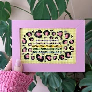 Puede incluir: Un bordado en punto de cruz enmarcado con un marco rosa. El bordado tiene un fondo amarillo con detalles de estampado de leopardo y el texto: "If you can't love yourself, how in the hell you gonna love somebody else?"