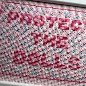 Puede incluir: Un bordado de punto de cruz enmarcado con las palabras "PROTECT THE DOLLS" en letras rosas. El fondo es de tela rosa claro con peque&ntilde;os detalles florales azules y blancos. El marco es blanco.