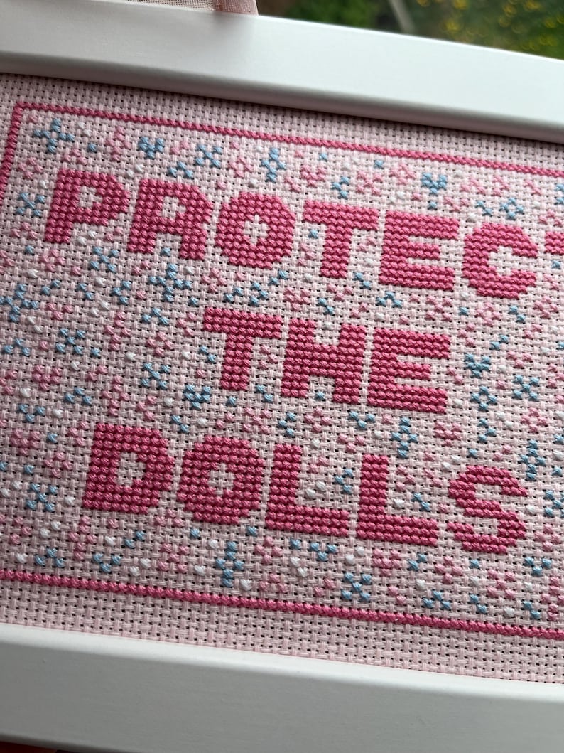 Puede incluir: Un bordado de punto de cruz enmarcado con las palabras "PROTECT THE DOLLS" en letras rosas. El fondo es de tela rosa claro con peque&ntilde;os detalles florales azules y blancos. El marco es blanco.
