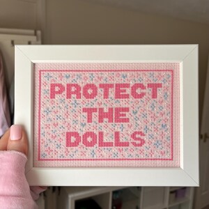 Puede incluir: Una obra de arte de punto de cruz enmarcada con las palabras "PROTECT THE DOLLS" en rosa. El fondo es un patr&oacute;n rosa y azul. El marco es blanco.