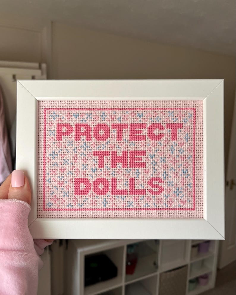 Puede incluir: Una obra de arte de punto de cruz enmarcada con las palabras "PROTECT THE DOLLS" en rosa. El fondo es un patr&oacute;n rosa y azul. El marco es blanco.
