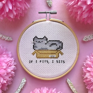 May include: Cross-stitch embroidery in a wooden hoop features a gray cat in a cardboard box. The text "IF I FITS, I SITS" is stitched below the cat. The background is pink, with pink flowers and white sprigs.