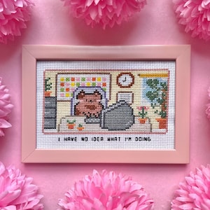 May include: Cross-stitch artwork in a pink frame. The cross-stitch depicts a cat at a desk with the text "I HAVE NO IDEA WHAT I'M DOING". The background is pink with pink paper flowers.