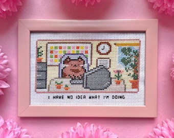 Könnte beinhalten: Kreuzstich-Kunstwerk in einem rosa Rahmen. Der Kreuzstich zeigt eine Katze an einem Schreibtisch mit dem Text "I HAVE NO IDEA WHAT I'M DOING". Der Hintergrund ist rosa mit rosa Papierblumen.