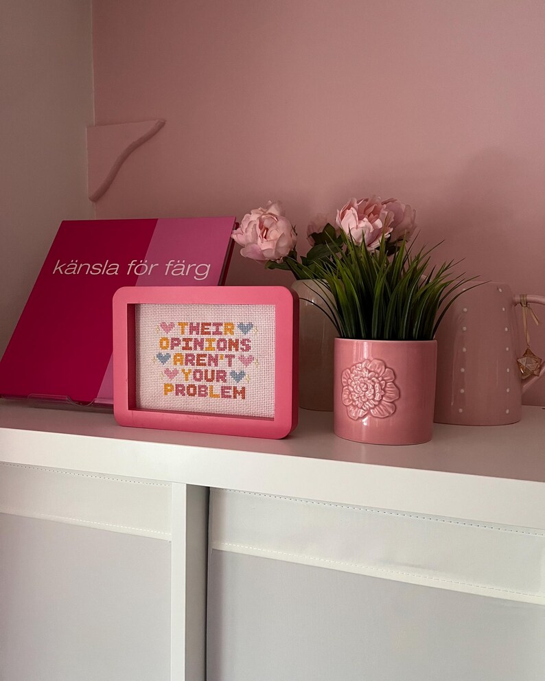 Puede incluir: Un bordado en punto de cruz enmarcado en rosa con el texto "THEIR OPINIONS AREN'T YOUR PROBLEM" descansa sobre una estanter&iacute;a blanca. Junto a &eacute;l, una maceta rosa con un dise&ntilde;o floral y un libro rosa con el texto "k&auml;nsla f&ouml;r f&auml;rg".