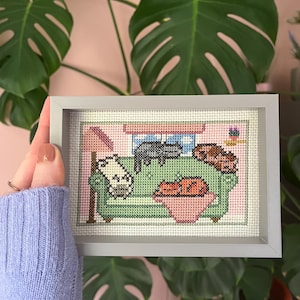 Może przedstawiać: Oprawiona praca haftu krzyżykowego przedstawia zieloną sofę z trzema kotami. Koty są w różnych pozach, jeden na sofie, jeden na podłokietniku i jeden w różowej misce. Rama jest szara.