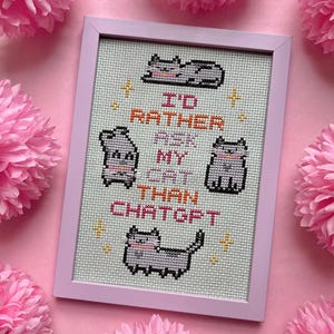 Pode incluir: Uma bordado em ponto cruz emoldurado, com uma moldura rosa claro, apresentando quatro ilustra&ccedil;&otilde;es de gatos cinzentos e o texto "I'D RATHER ASK MY CAT THAN CHATGPT" em laranja e preto. O fundo &eacute; tecido verde claro. Flores de papel rosa circundam a moldura.