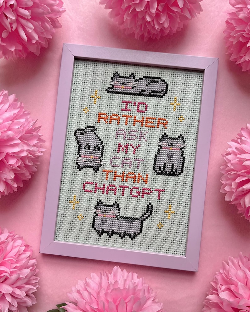 Pode incluir: Uma bordado em ponto cruz emoldurado, com uma moldura rosa claro, apresentando quatro ilustra&ccedil;&otilde;es de gatos cinzentos e o texto "I'D RATHER ASK MY CAT THAN CHATGPT" em laranja e preto. O fundo &eacute; tecido verde claro. Flores de papel rosa circundam a moldura.