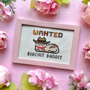 Puede incluir: Un bordado de punto de cruz enmarcado con un gato de dibujos animados con sombrero de vaquero, con el texto "WANTED BISCUIT BANDIT". El marco es rosa claro, sobre un fondo rosa claro con flores rosas.