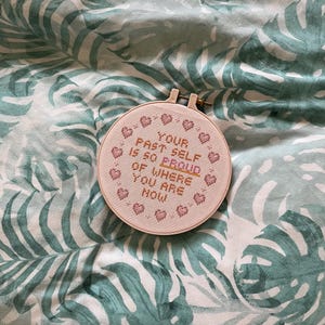 K&ouml;nnte beinhalten: Ein runder Kreuzstich-Stickrahmen mit dem Text "YOUR PAST SELF IS SO PROUD OF WHERE YOU ARE NOW" in rosa Garn. Das Design enth&auml;lt kleine Herzmotive. Der Rahmen befindet sich vor einem Hintergrund mit einem gr&uuml;nen und wei&szlig;en tropischen Bl&auml;ttermuster.