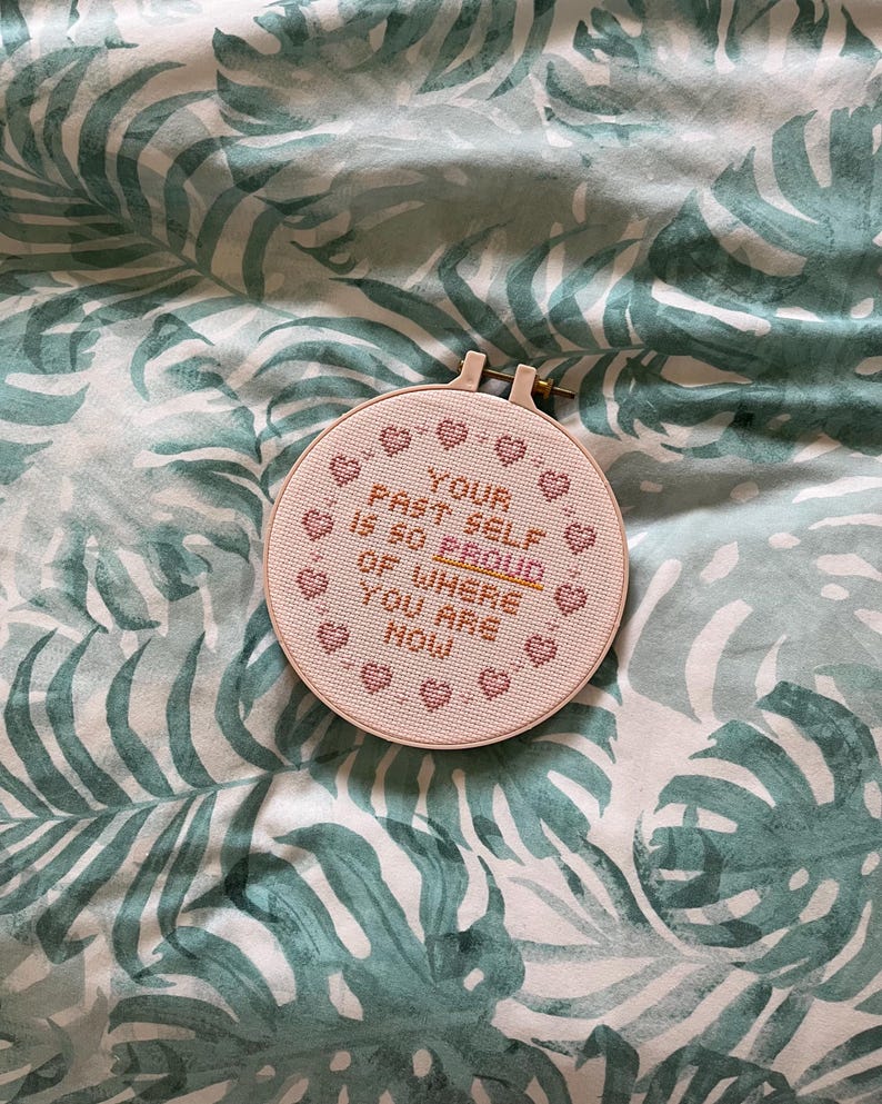 K&ouml;nnte beinhalten: Ein runder Kreuzstich-Stickrahmen mit dem Text "YOUR PAST SELF IS SO PROUD OF WHERE YOU ARE NOW" in rosa Garn. Das Design enth&auml;lt kleine Herzmotive. Der Rahmen befindet sich vor einem Hintergrund mit einem gr&uuml;nen und wei&szlig;en tropischen Bl&auml;ttermuster.