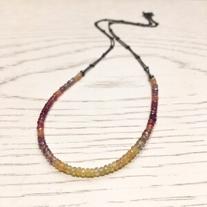 May include: A delicate necklace with a gradient of gemstone beads, ranging from deep red to yellow. The beads are strung on a black chain.