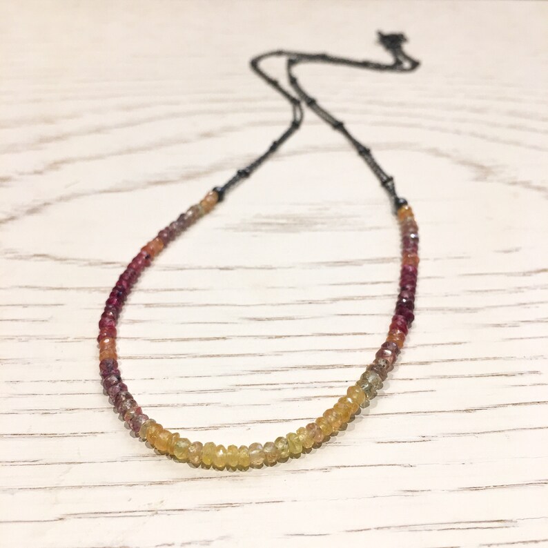 May include: A delicate necklace with a gradient of gemstone beads, ranging from deep red to yellow. The beads are strung on a black chain.