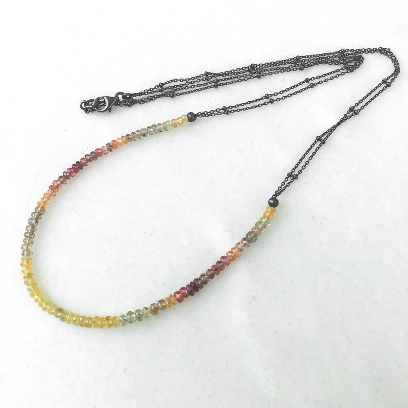 May include: A multi-colored gemstone necklace with a graduated color scheme of red, orange, yellow, and green. The necklace is strung on a black chain with a lobster clasp.