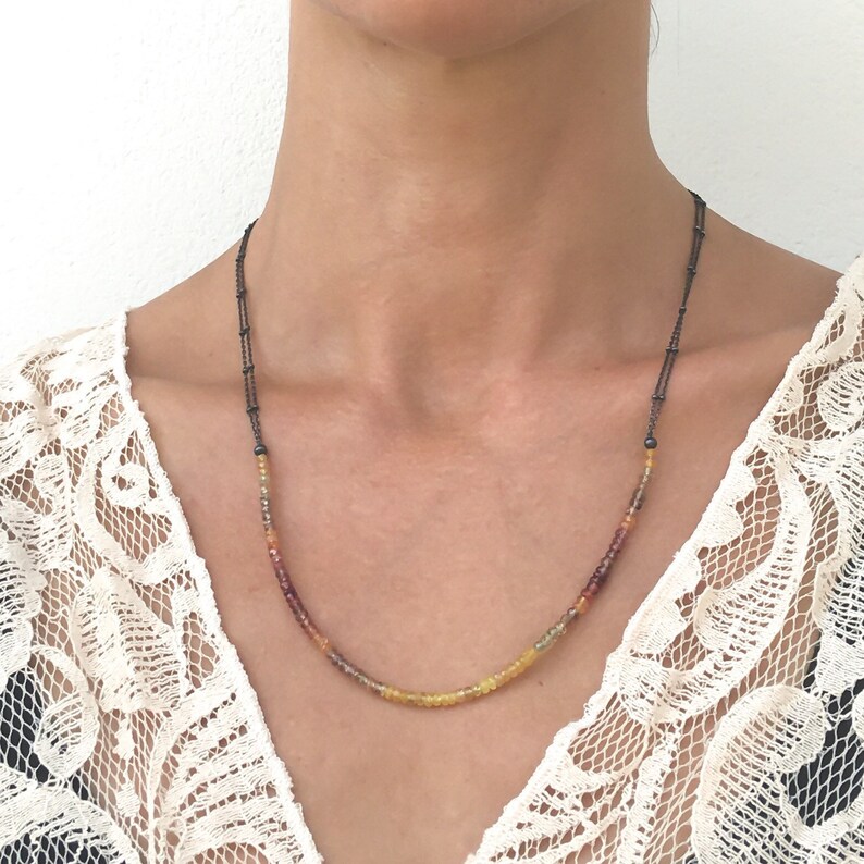 May include: A delicate necklace with a graduated ombre design of small, round, gemstone beads in shades of red, orange, and yellow. The necklace is strung on a thin, black cord.