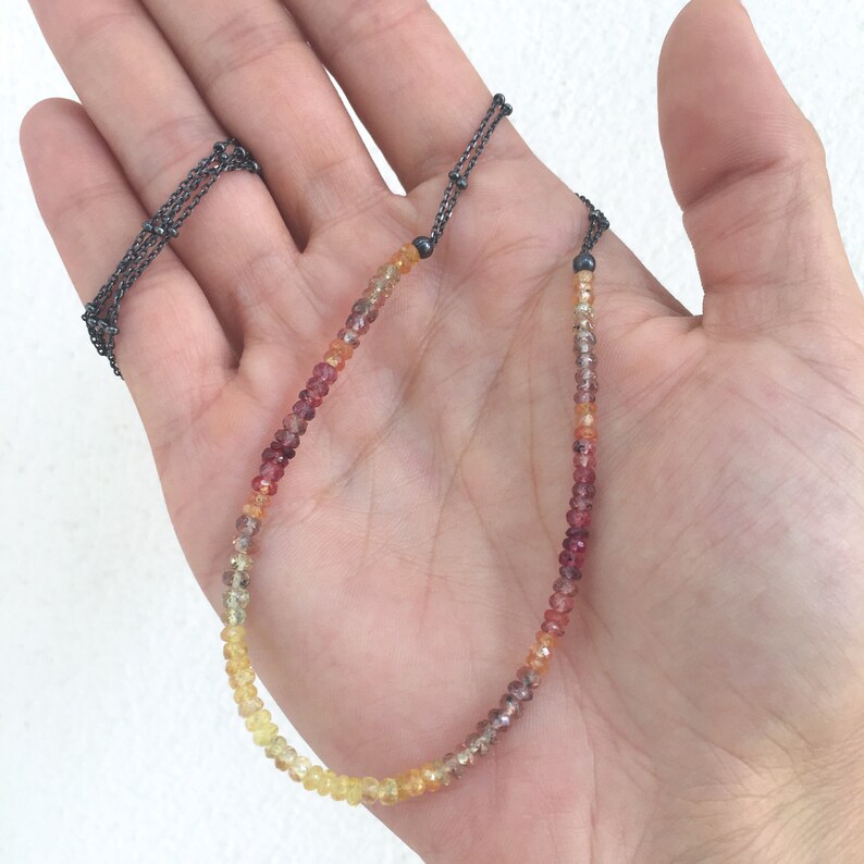 May include: A delicate necklace with a graduated ombre design of yellow, orange, and red gemstones. The necklace is made with a silver chain and black beads.