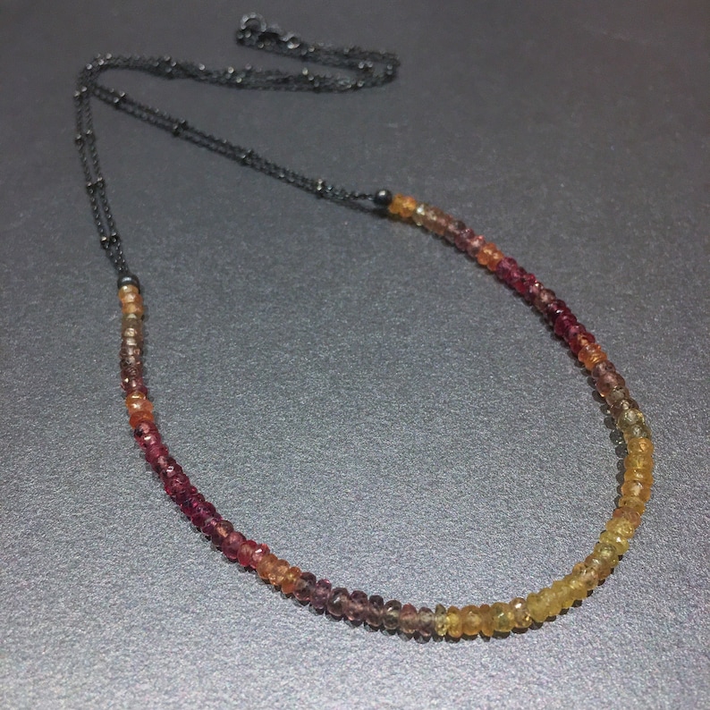 May include: A delicate necklace with a graduated ombre of red, orange, and yellow gemstones. The necklace is made with a black chain and features a simple clasp.