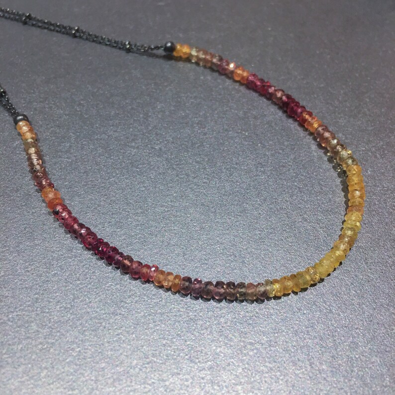 May include: A multi-colored gemstone necklace with a gradient of red, orange, and yellow beads. The necklace is made with a black cord.