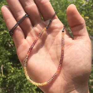 May include: A delicate necklace with a graduated ombre effect of yellow, orange, and red gemstones. The necklace is made with a thin black chain.