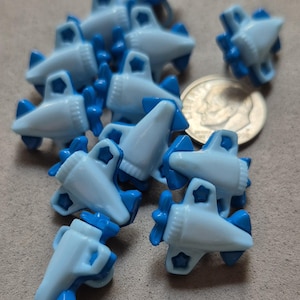 May include: A collection of light blue airplane-shaped charms with dark blue accents, including propellers and tail fins. The charms are scattered on a neutral-toned surface, with a U.S. dime for size comparison. These charms are likely for crafting or jewelry making.