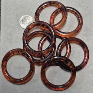 May include: A collection of translucent, tortoiseshell-patterned rings. The rings are a mix of brown and amber hues, with a marbled effect. A U.S. dime is included for size comparison. The rings are likely for jewelry or craft projects.